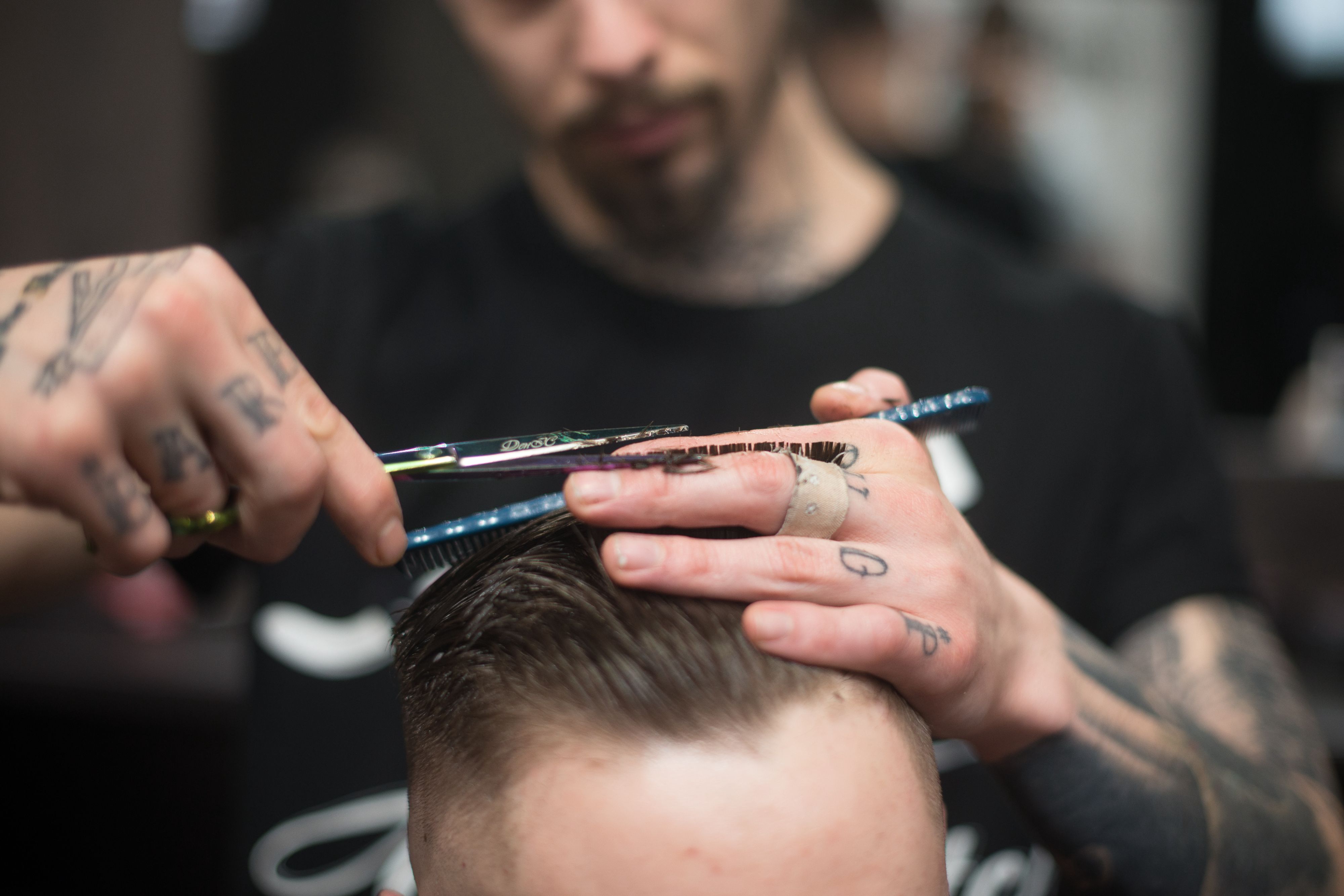 A barber with tattoos on his finger cutting a guy hair.